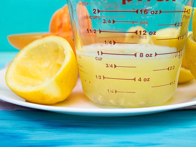A cup of lemon juice in a glass measuring cup next to lemon halves 